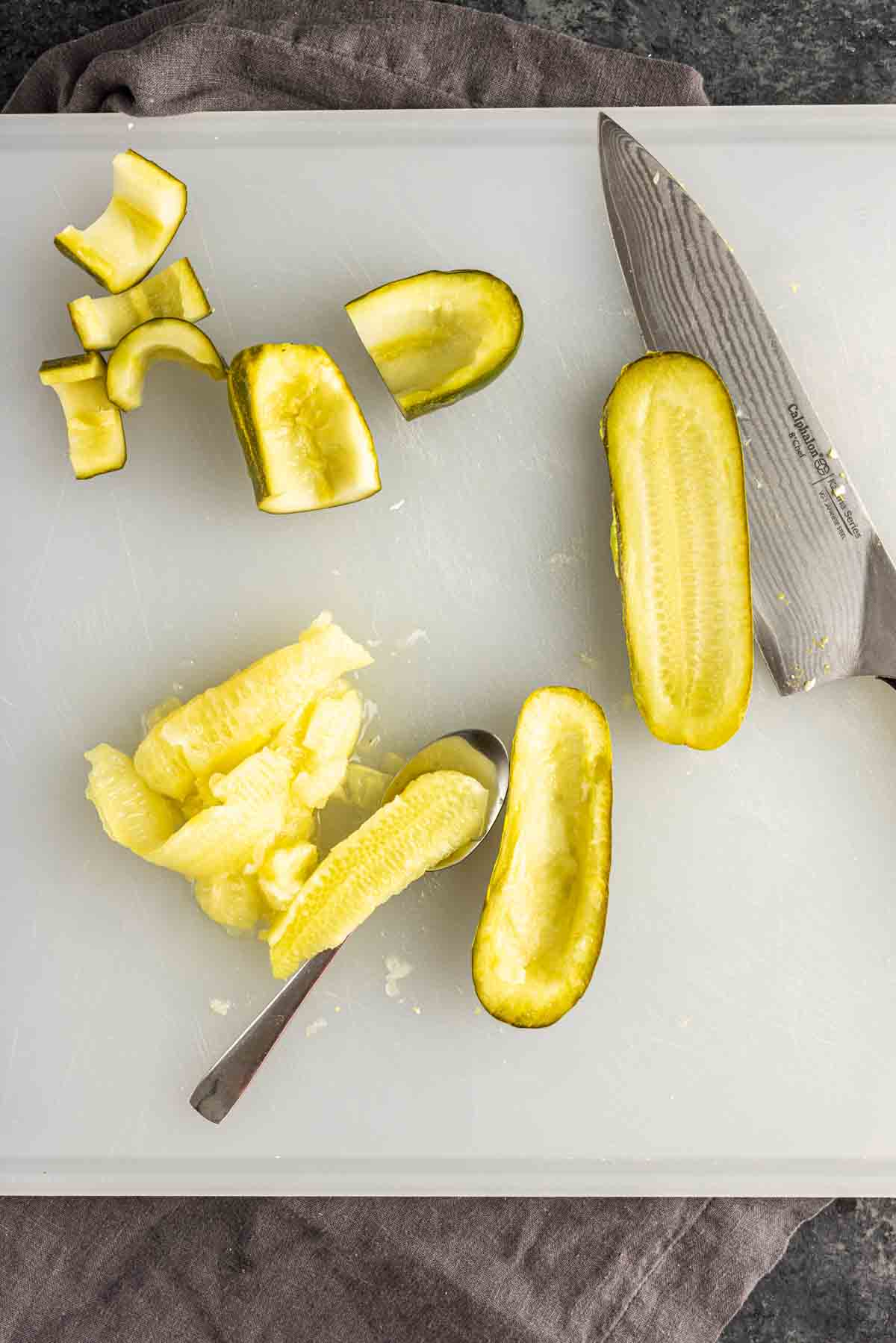 A cutting board with a knife, a halved pickle being hollowed out by a spoon, and pieces of sliced pickles with scooped-out insides—perfect for making deviled pickle recipe appetizers or unique twists on deviled eggs with pickles.