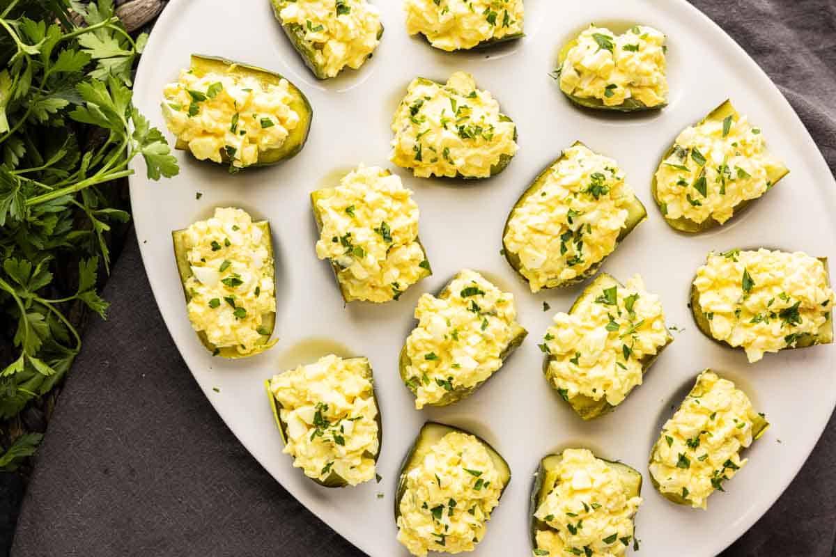 A white plate with several halved green bell peppers filled with egg salad, resembling easy pickle appetizers, garnished with chopped herbs, on a dark surface next to fresh parsley.