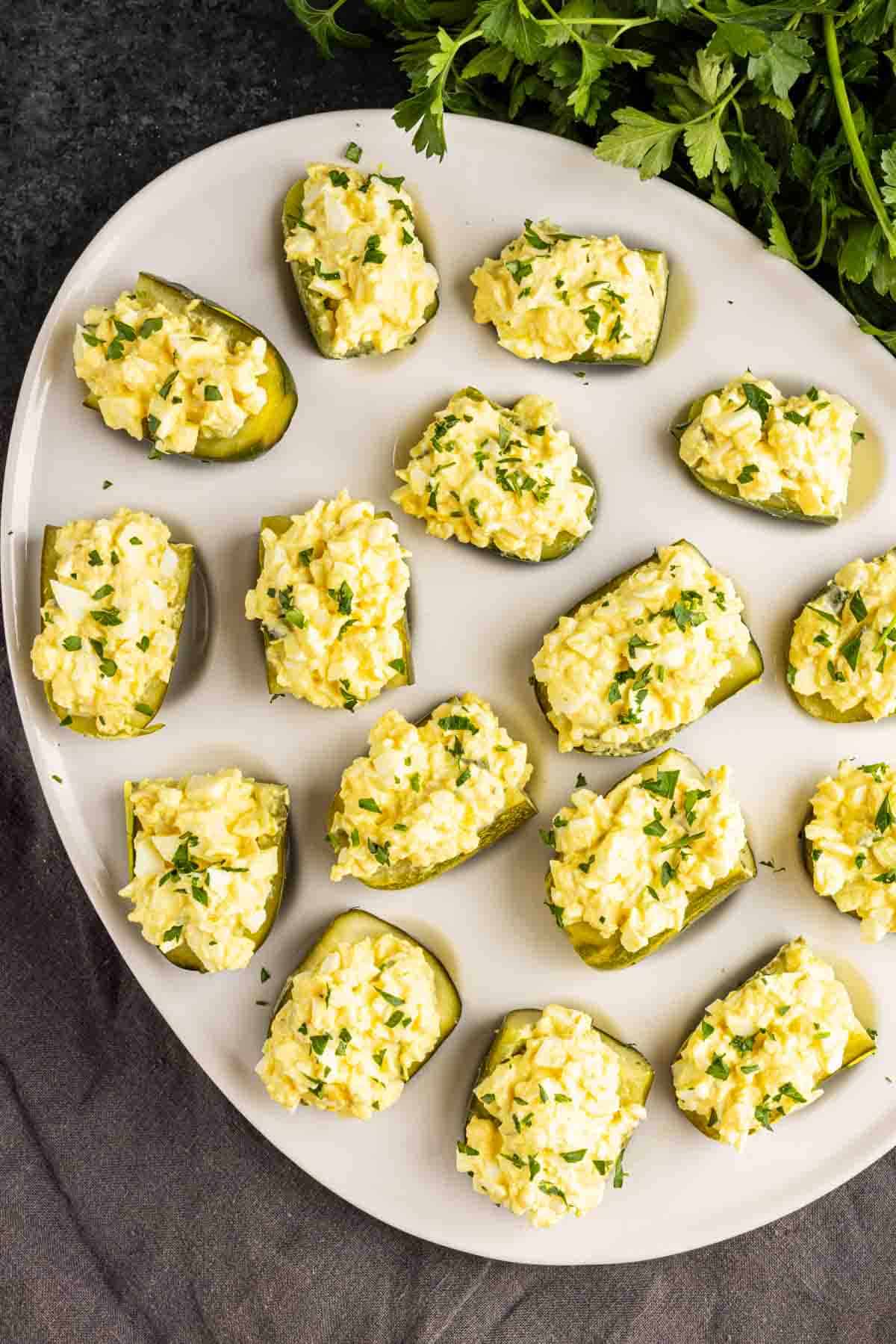 A white plate with Deviled Pickles—pickle halves filled with egg salad, garnished with chopped herbs, on a dark surface with fresh parsley nearby. This tasty deviled pickle recipe is perfect for easy pickle appetizers.