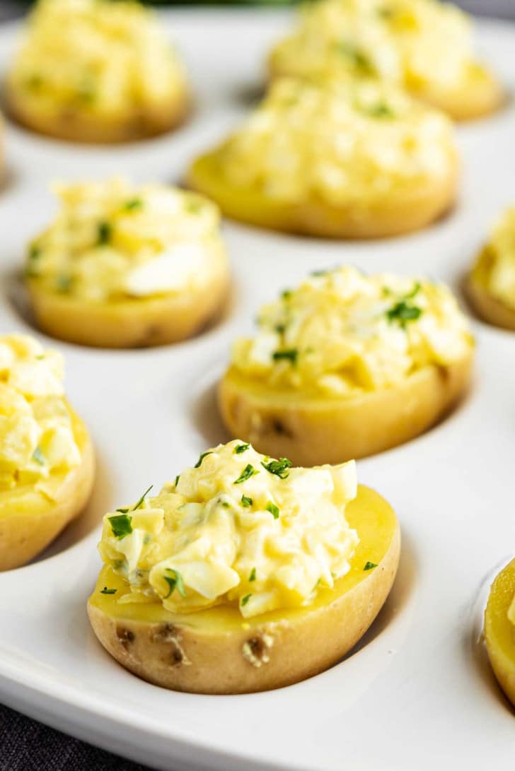 Halved baby potatoes topped with a creamy egg salad mixture and garnished with chopped herbs, arranged on a white serving tray.