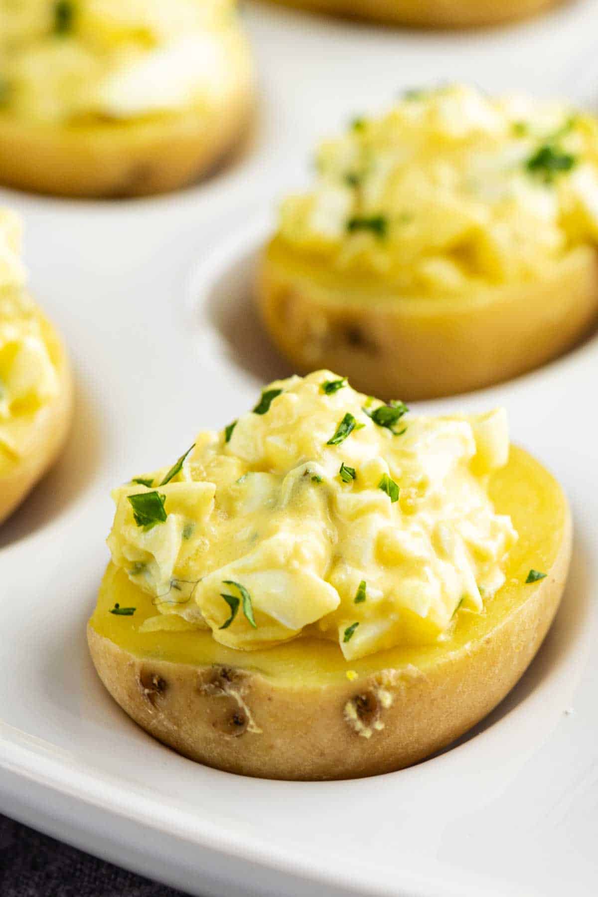 Halved small potatoes topped with creamy egg salad and parsley, these party potato bites are arranged on a white serving tray—an irresistible deviled potato recipe perfect for any gathering.