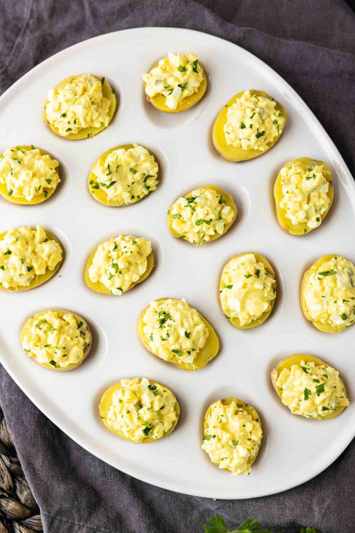 A white platter with halved small potatoes topped with a creamy, yellow egg salad mixture and garnished with chopped herbs.