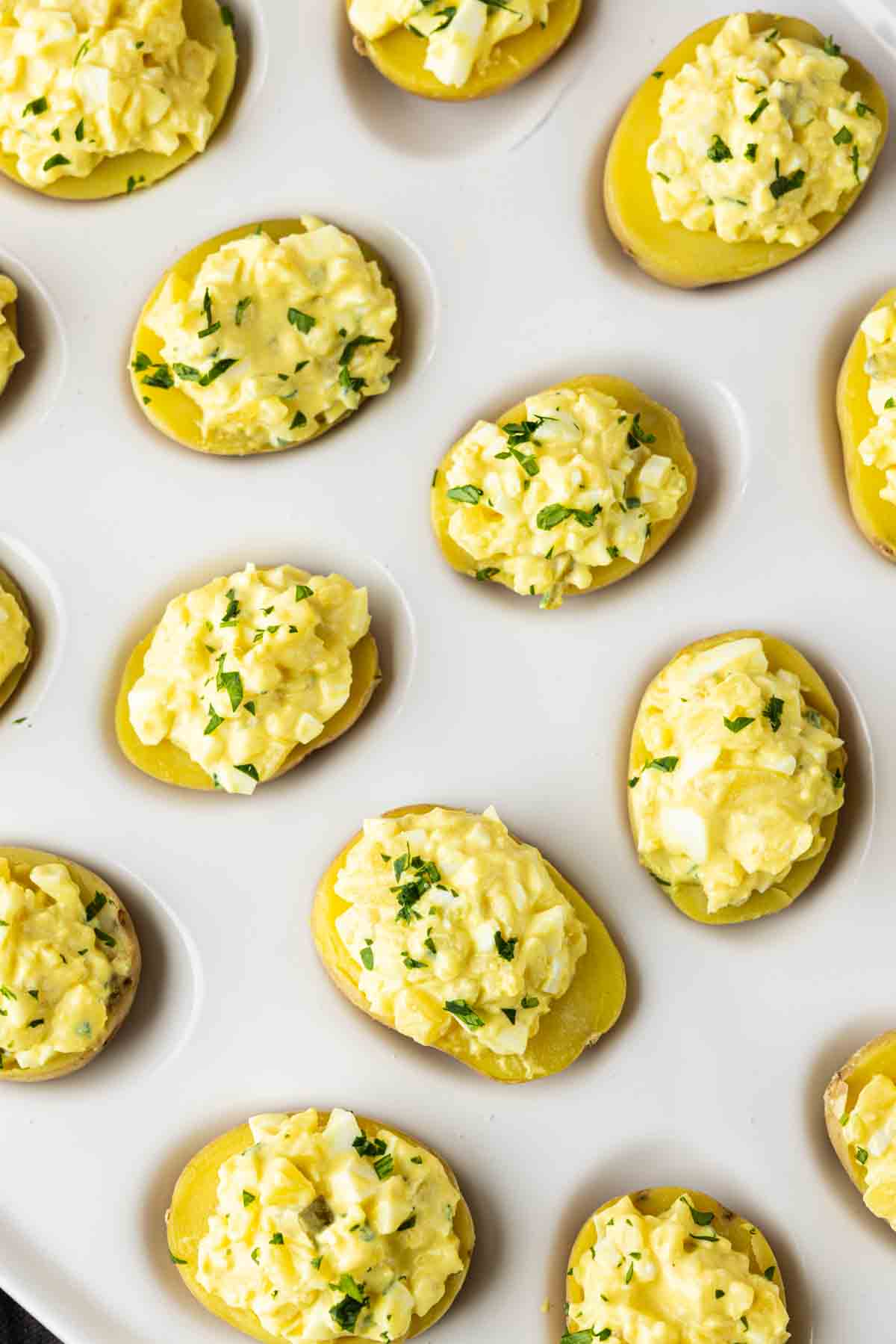 Halved potatoes topped with a yellow egg salad mixture and chopped herbs, these party potato bites make an easy potato appetizer, perfectly arranged on a white surface.