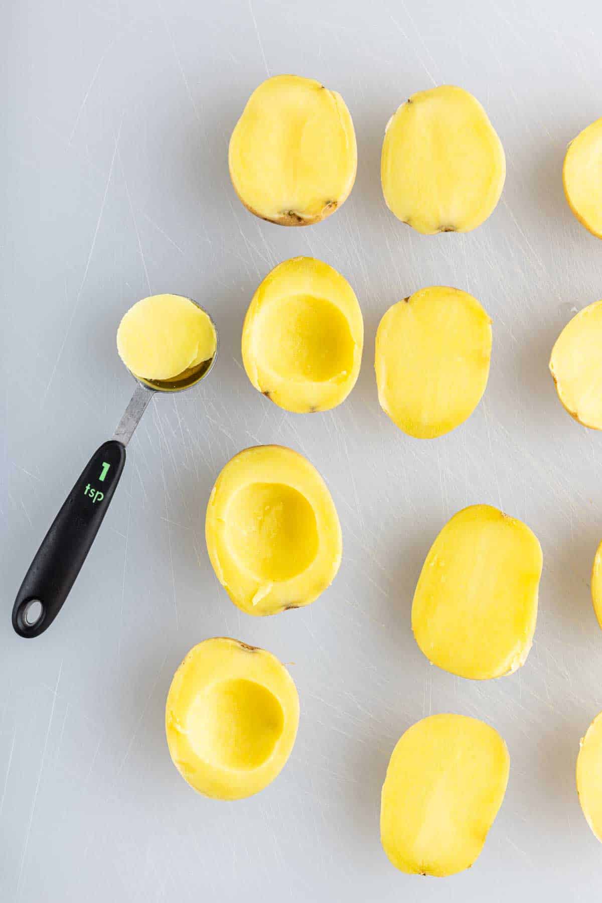 Several yellow potato halves are shown on a white surface, some hollowed out with a melon baller labeled "1 tsp," ready to be filled for a tasty deviled potato recipe or served as party potato bites.