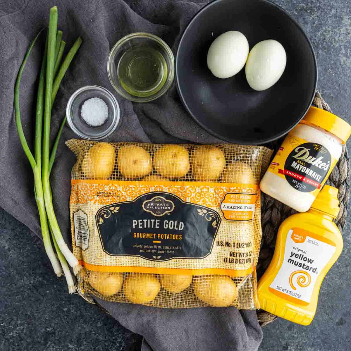 A bag of petite gold potatoes, two boiled eggs in a bowl, green onions, mayo, mustard, salt, and oil—everything you need for deviled potatoes or an easy potato appetizer perfect for party potato bites on a gray cloth.