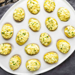 A white platter with several deviled eggs topped with a chopped egg mixture and garnished with green herbs.