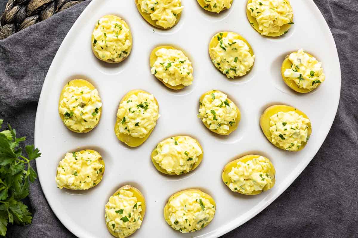 A white oval platter displays halved potatoes filled with a creamy, yellow egg salad mixture, garnished with chopped herbs—perfect party potato bites inspired by a classic deviled potato recipe.