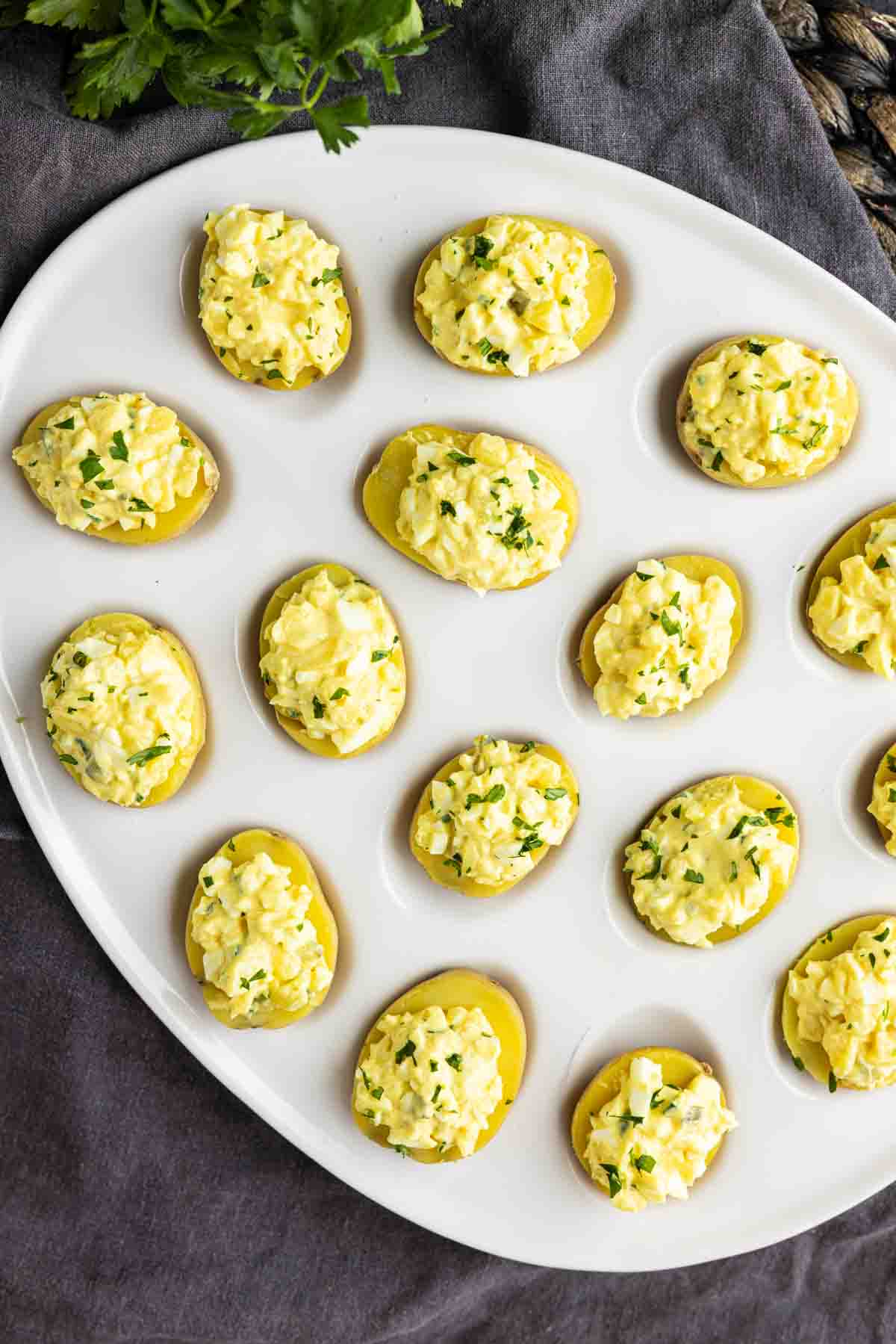 A white plate with several halved boiled potatoes topped with a yellow egg salad mixture and garnished with chopped herbs, making these party potato bites an easy potato appetizer for any gathering.