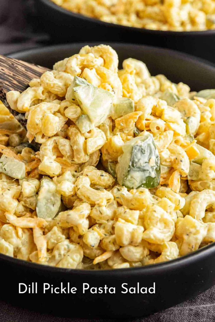 A bowl of dill pickle pasta salad with elbow macaroni, chopped pickles, and a creamy dressing, served with a wooden spoon.