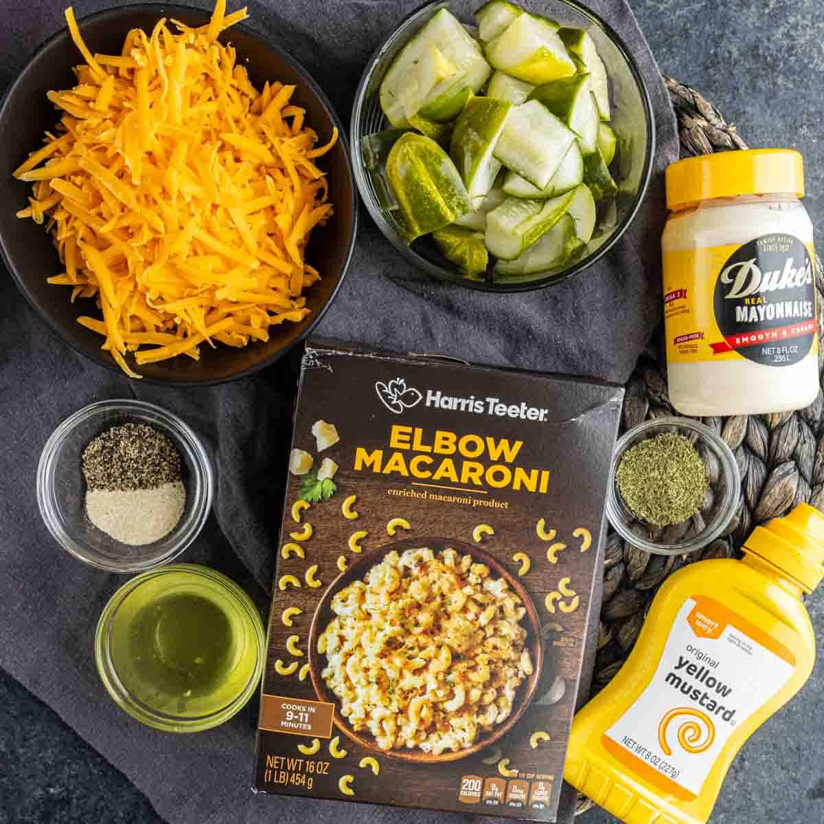 Ingredients for an easy pasta salad recipe arranged on a surface: shredded cheddar, elbow macaroni, chopped pickles, mayonnaise, yellow mustard, oil, pepper, herbs—all ready for a tangy dill pickle juice dressing.