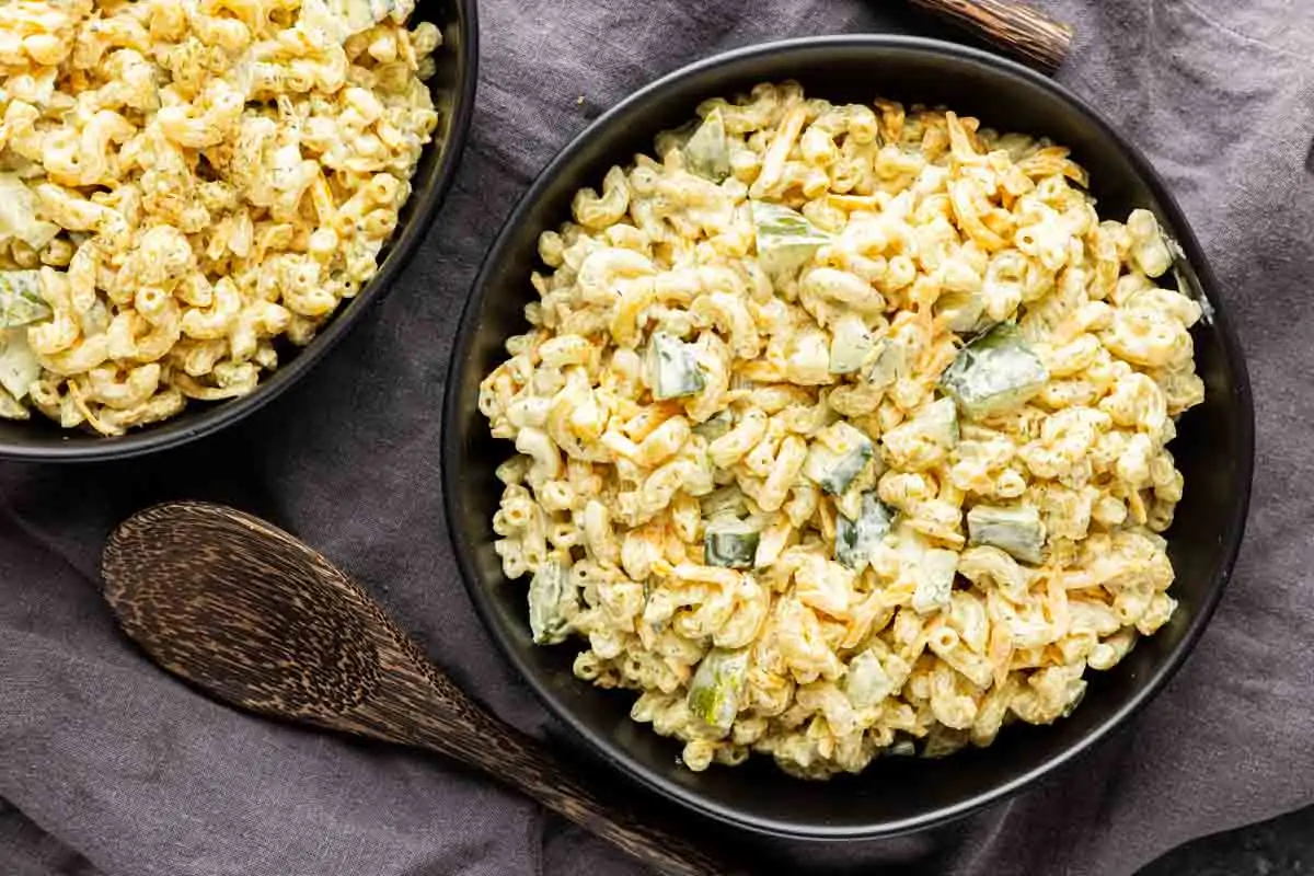 Two black bowls filled with Dill Pickle Pasta Salad—elbow pasta, diced vegetables, pickles, and a creamy dressing—rest on a dark cloth with wooden serving utensils nearby. An easy pasta salad recipe for any occasion.