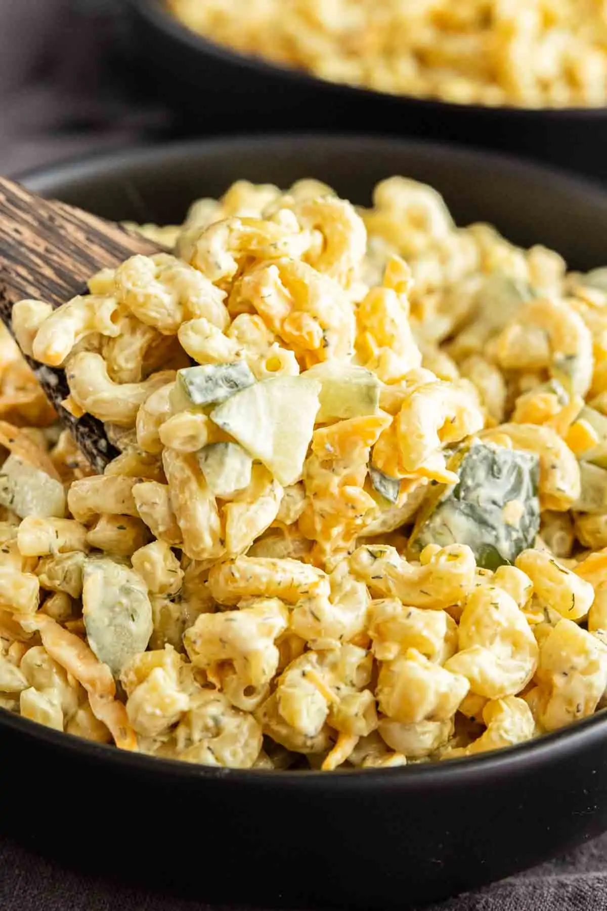 A close-up of Dill Pickle Pasta Salad with chopped pickles and a creamy dill pickle juice dressing, being stirred with a wooden spoon in a black bowl.