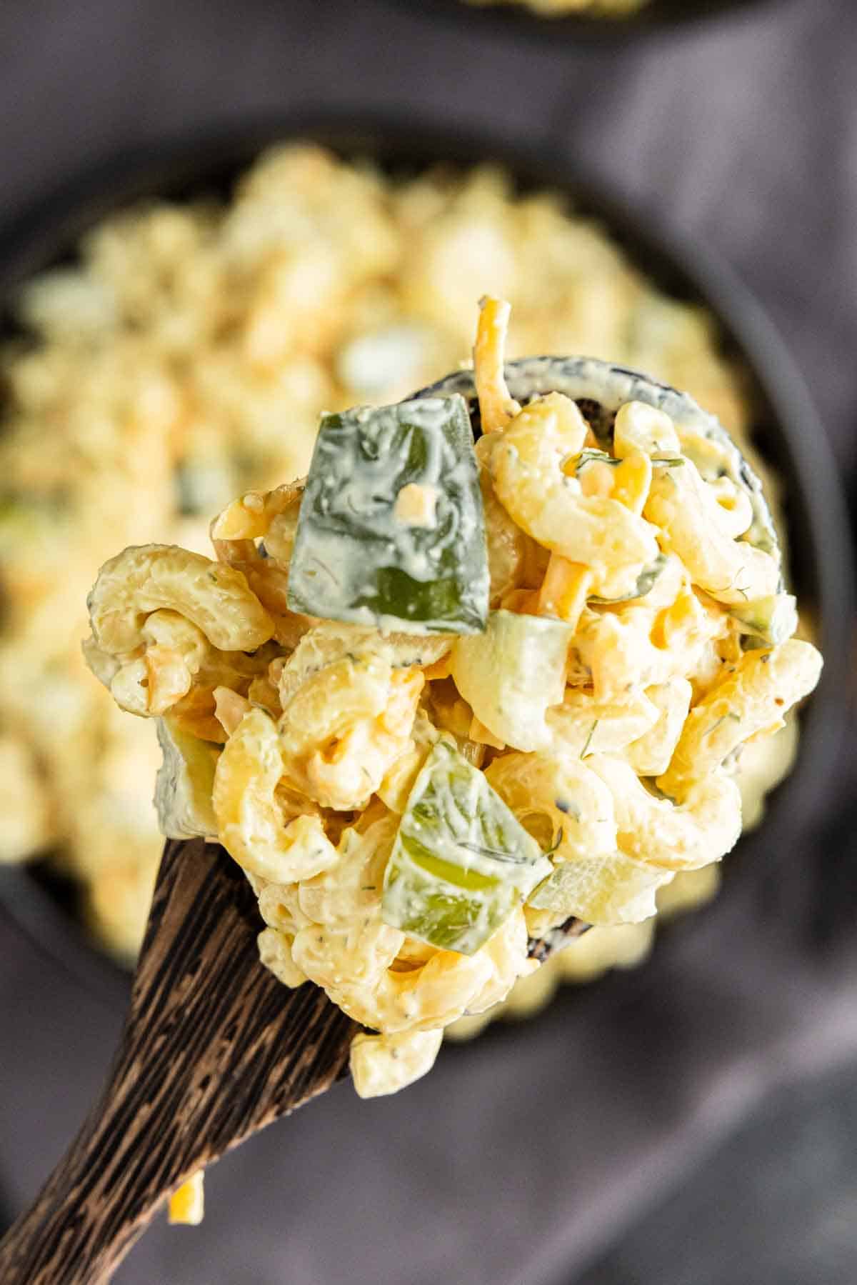 A close-up of a wooden spoon holding creamy pasta salad with diced green peppers and dill pickle juice dressing, above a bowl of more salad.
