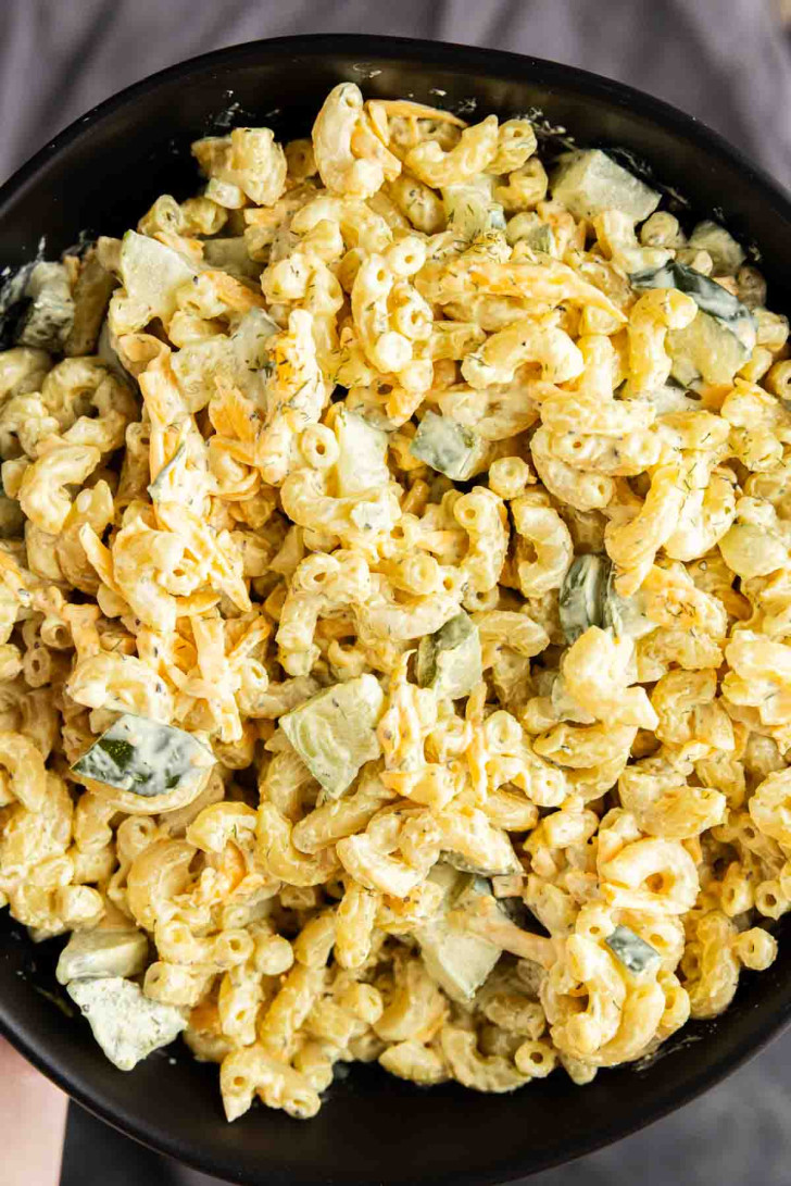 A close-up of macaroni salad with diced cucumbers, dressed in a creamy yellow sauce, served in a black bowl.
