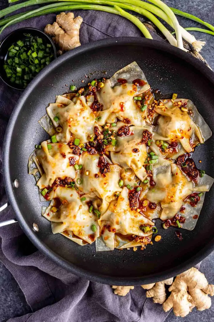 Pan of cooked dumplings topped with chili oil and chopped green onions, surrounded by fresh ginger, scallions, and a bowl of chopped herbs.