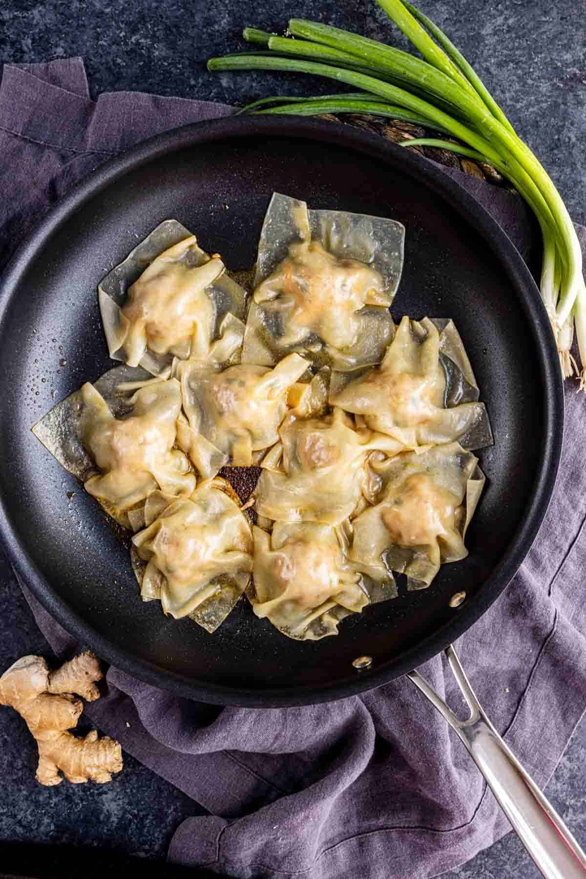 A black skillet with eight golden pork dumplings, inspired by viral TikTok dumplings, sits on a dark cloth, with fresh ginger and green onions nearby.