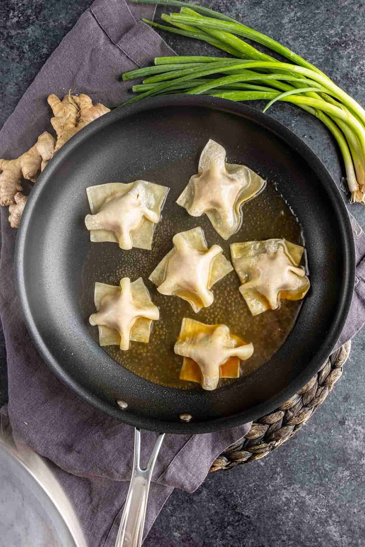 Six pork dumplings are being pan-fried in oil in a black skillet; ginger root and green onions are placed nearby on a dark countertop.