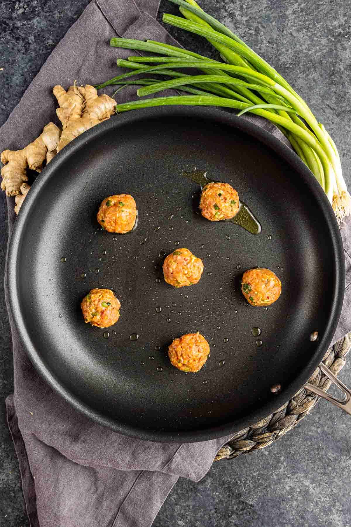 Six uncooked meatballs in a black skillet with a bit of oil, placed on a dark cloth next to fresh ginger and green onions—perfect for making viral blanket dumplings or wonton wrapper dumplings on a dark textured surface.