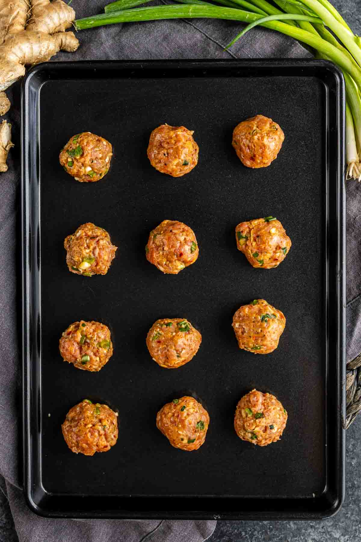 Twelve uncooked pork dumplings with herbs are evenly spaced on a black baking sheet, with fresh ginger and green onions visible in the background—perfect for making no fold dumplings or wonton wrapper dumplings.