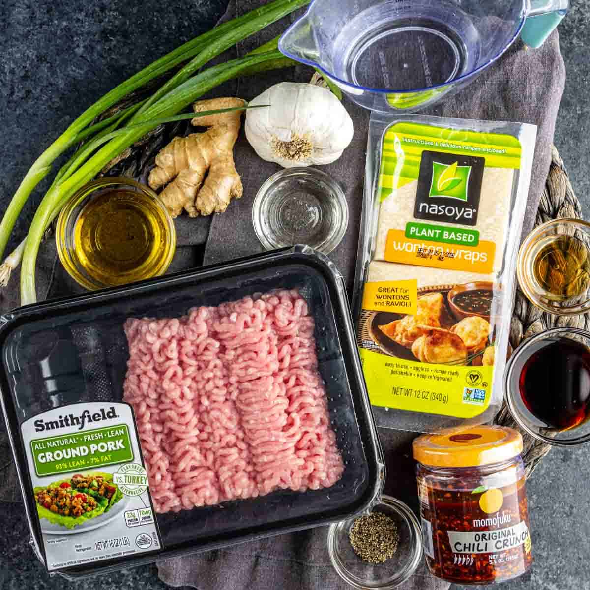 Various ingredients are arranged on a table, including ground pork, wonton wraps for TikTok dumplings, chili crunch, green onions, ginger, garlic, seasonings, and a measuring cup with water.