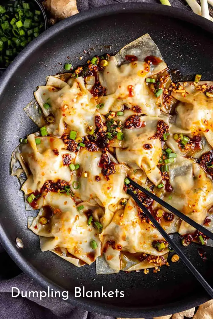 A pan filled with cooked dumplings topped with chili oil and chopped green onions, with chopsticks picking up one dumpling. Text reads "Dumpling Blankets.