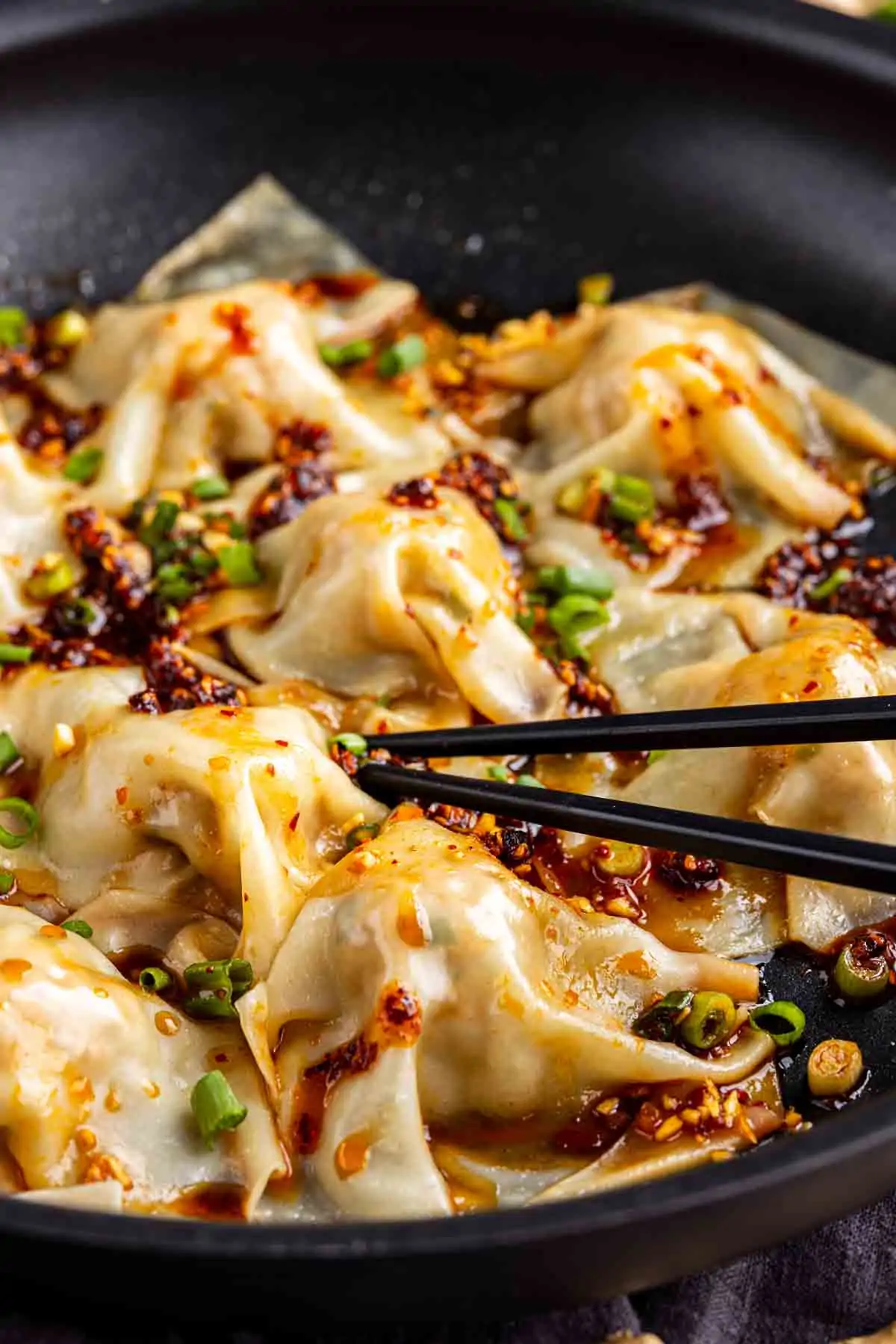 A close-up of no fold dumplings, topped with chili oil and chopped green onions in a black pan, with a pair of chopsticks picking up one TikTok dumpling.