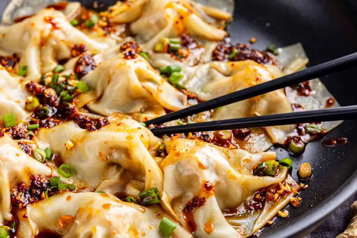 Black chopsticks hold a pork dumpling topped with chili oil and chopped green onions, with several more no fold dumplings in sauce visible in a skillet.