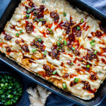 Rectangular baking dish with layered viral dumpling lasagna topped with spicy chili oil and chopped green onions, next to a bowl of extra green onions and a piece of ginger.