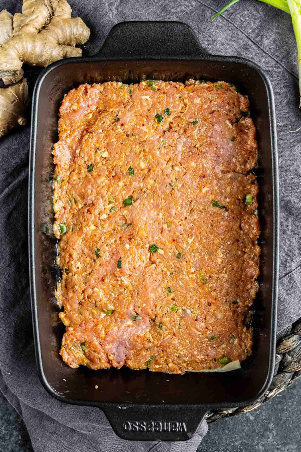 A rectangular black baking dish filled with seasoned, uncooked ground meat mixture for a pork dumpling casserole, placed on a dark cloth next to ginger root and green onions.