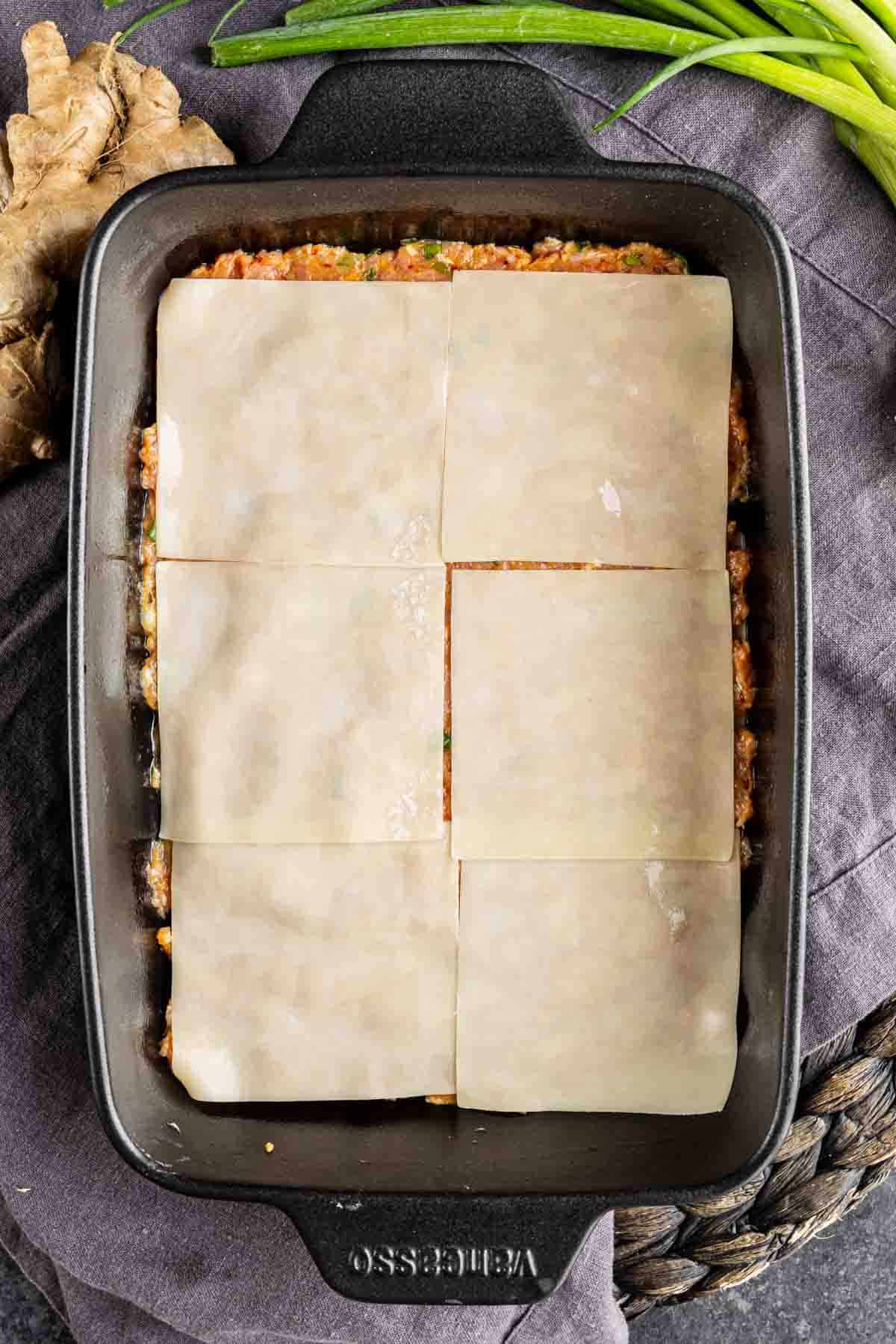 A black rectangular baking dish filled with layered noodles and filling, topped with six uncooked lasagna sheets, resembles a creative dumpling lasagna. Green onions and ginger sit beside the dish, perfect for a TikTok dumpling lasagna recipe.
