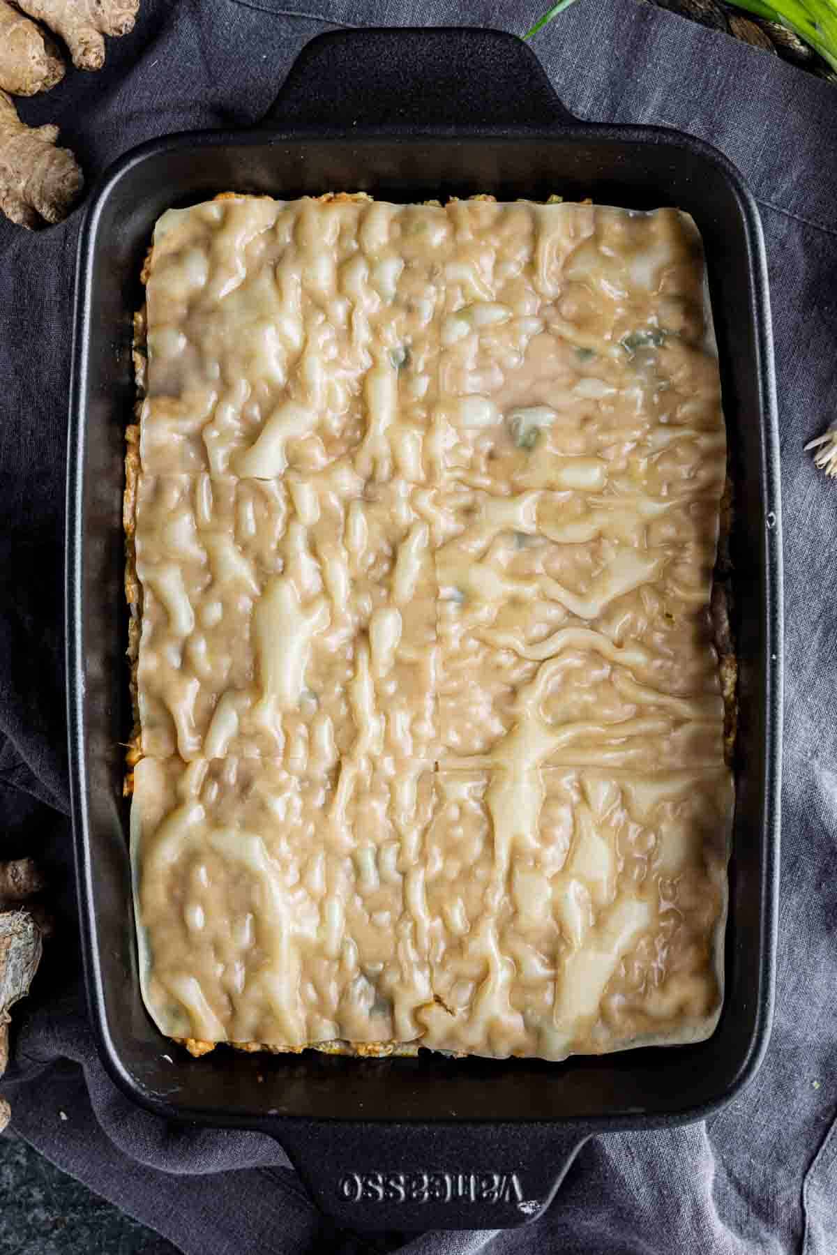 Rectangular baking dish with an unbaked viral dumpling lasagna topped with an even layer of creamy sauce, placed on a dark cloth.