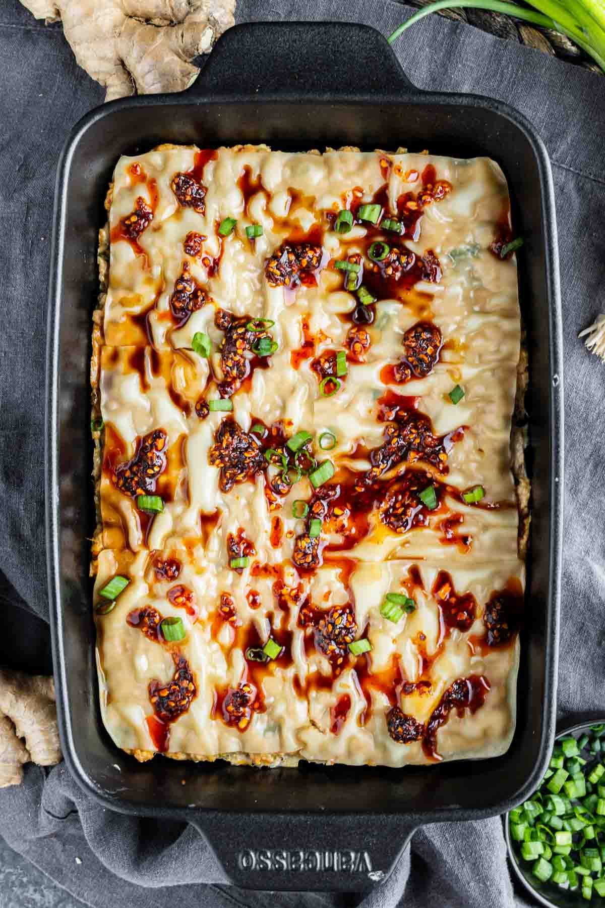Rectangular baking dish filled with a pork dumpling casserole topped with sauces, chopped green onions, and chili flakes, placed on a dark cloth.