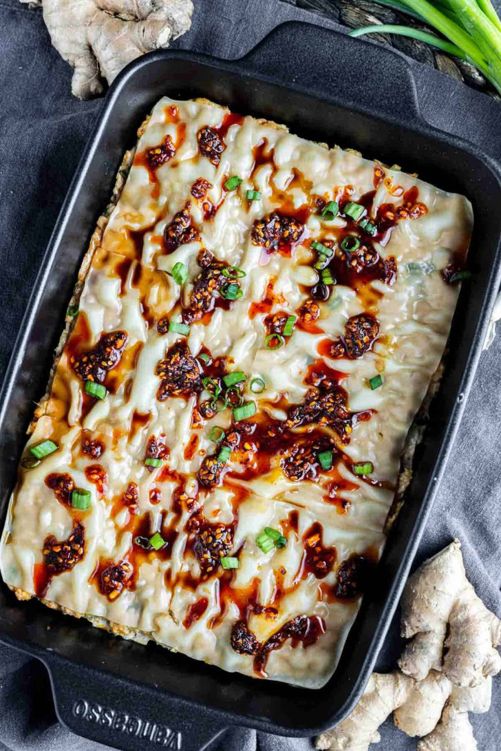 A rectangular baking dish with a baked noodle casserole topped with chili oil, green onions, and sauce, set on a dark background with ginger and green onions nearby.