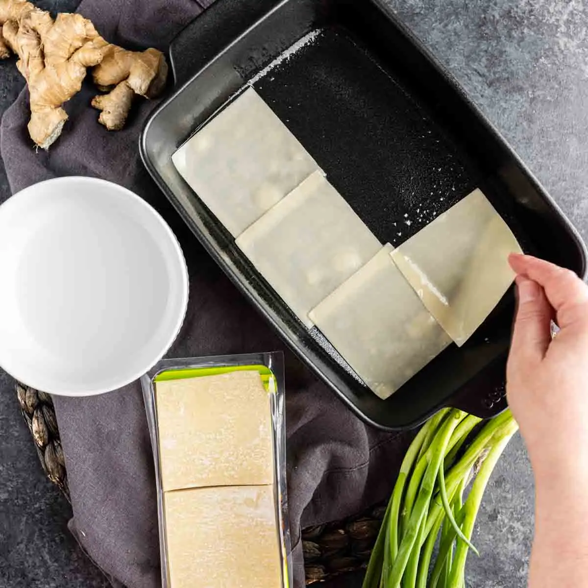 A hand places a wonton wrapper into a baking dish for a pork dumpling casserole; nearby are green onions, ginger, a bowl of water, and a pack of wonton wrappers—a nod to the viral dumpling lasagna trend.