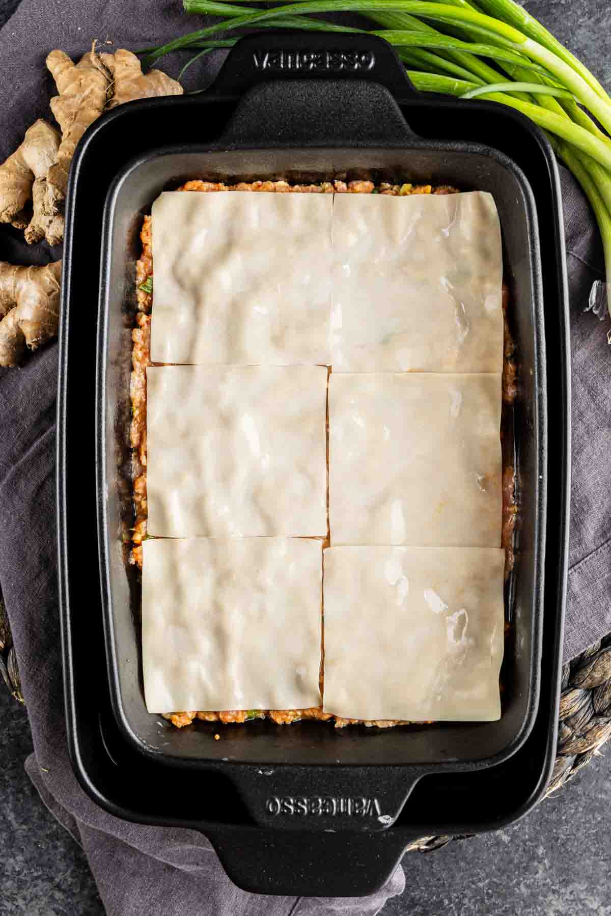 A rectangular baking dish filled with a layered casserole, topped with six sheets of uncooked pasta, hints at a viral dumpling lasagna inspired by TikTok. Green onions and ginger root are visible nearby.
