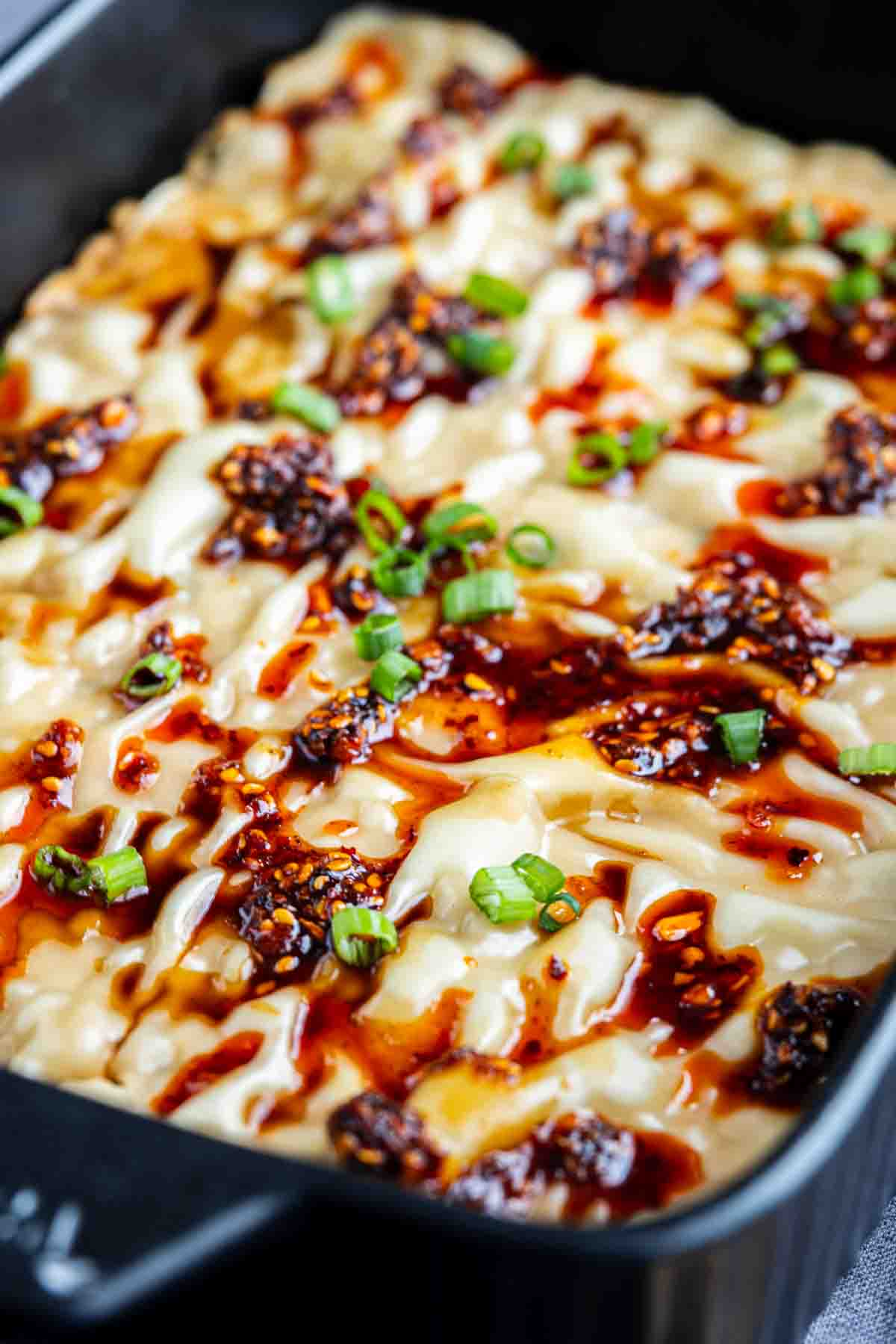 A close-up of a baked TikTok dumpling lasagna topped with melted cheese, chili oil, and chopped green onions in a black baking dish.