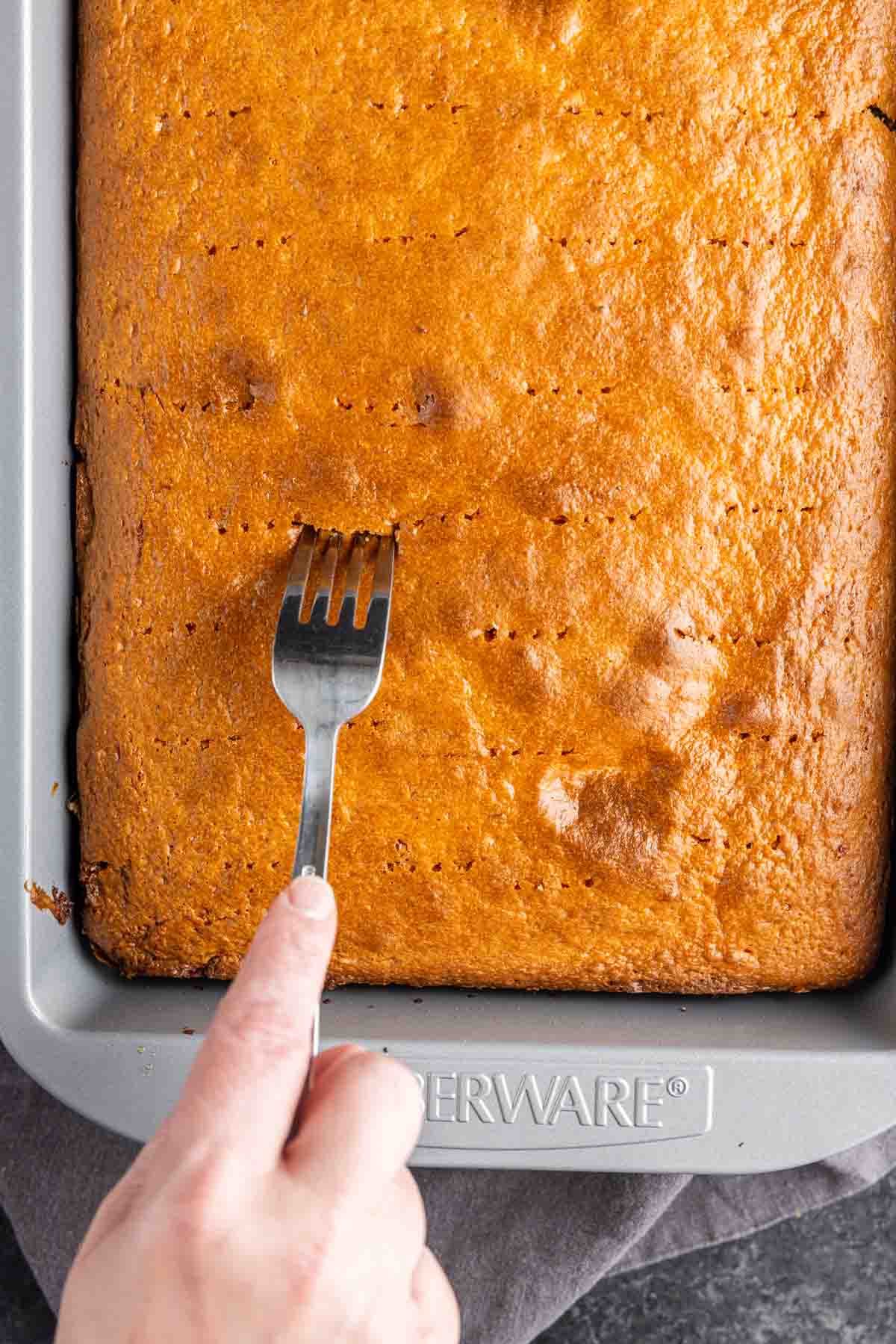 A hand uses a fork to poke holes in a golden-brown Honey Bun Cake in a rectangular baking pan.