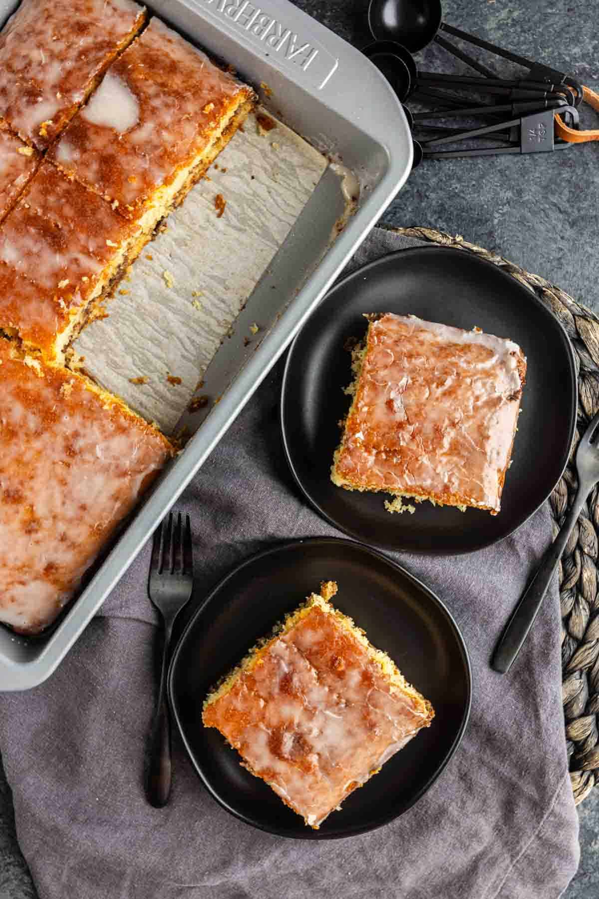 A baking pan with cinnamon swirl cake, some pieces removed, sits beside two black plates each holding a square piece of the cake. Black utensils and measuring spoons are nearby.