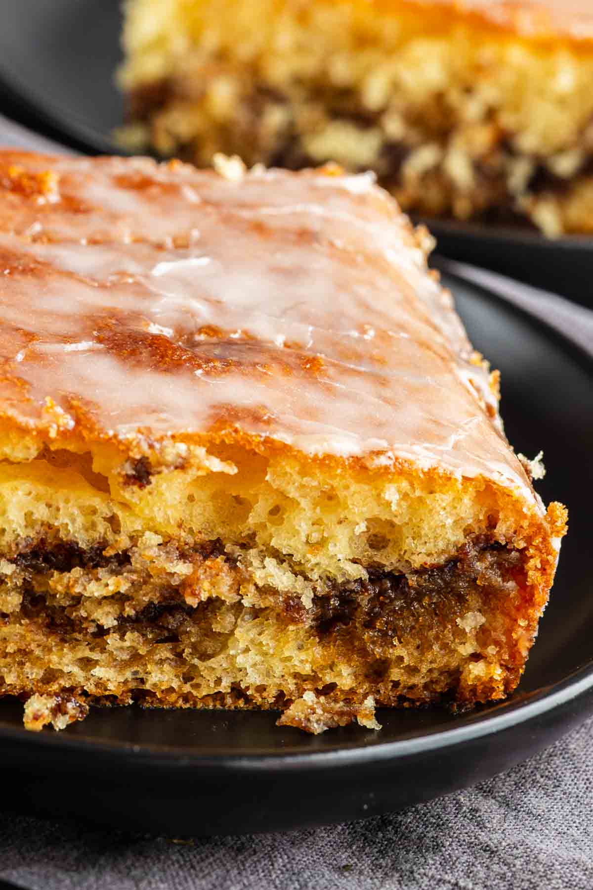 A close-up of a slice of Honey Bun Cake with a visible cinnamon swirl, glazed and served on a black plate.