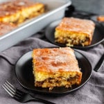 A slice of cinnamon swirl cake, made from an easy honey bun cake recipe and topped with sweet glaze, is served on a black plate, with a pan of more cake in the background.