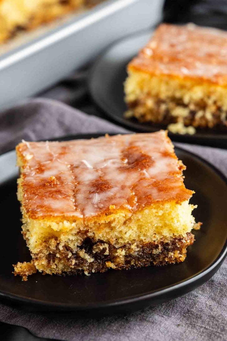 A square slice of glazed coffee cake with a crumbly, layered texture is served on a black plate.