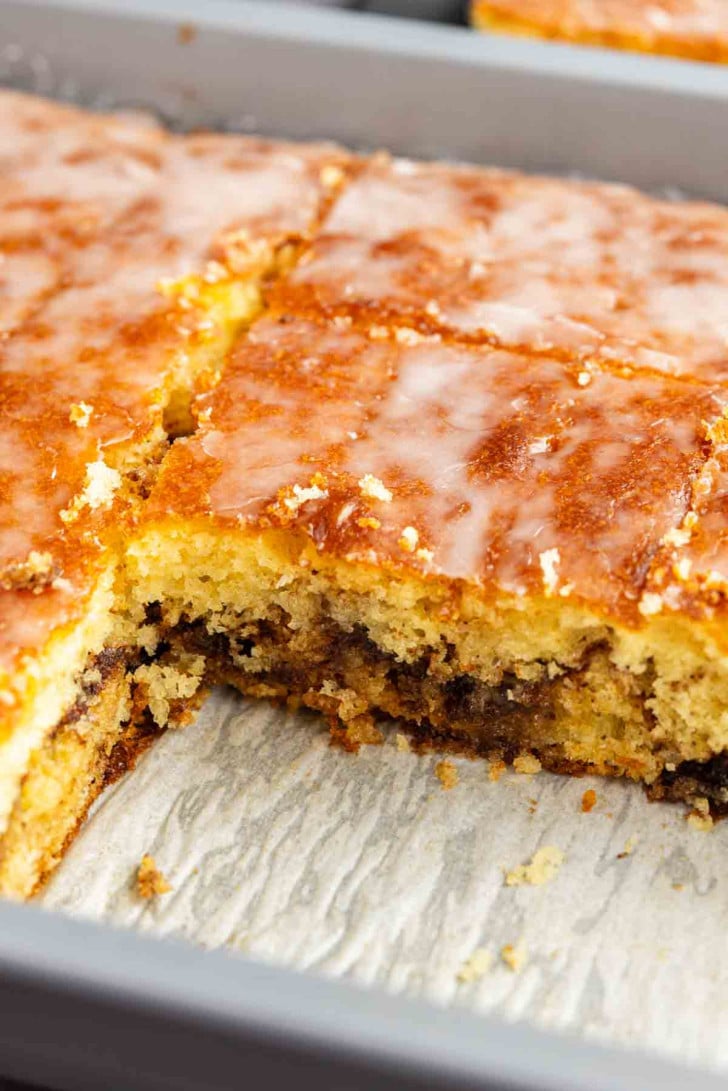 A close-up of sliced coffee cake with a cinnamon swirl and a glaze topping in a baking tray, with one piece partially removed.