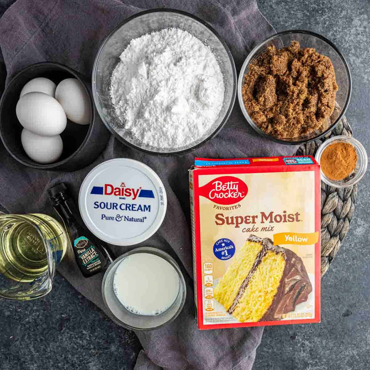 Ingredients for a Honey Bun Cake laid out on a table, including eggs, powdered sugar, brown sugar, cinnamon, cake mix, vanilla extract, oil, milk, and sour cream.