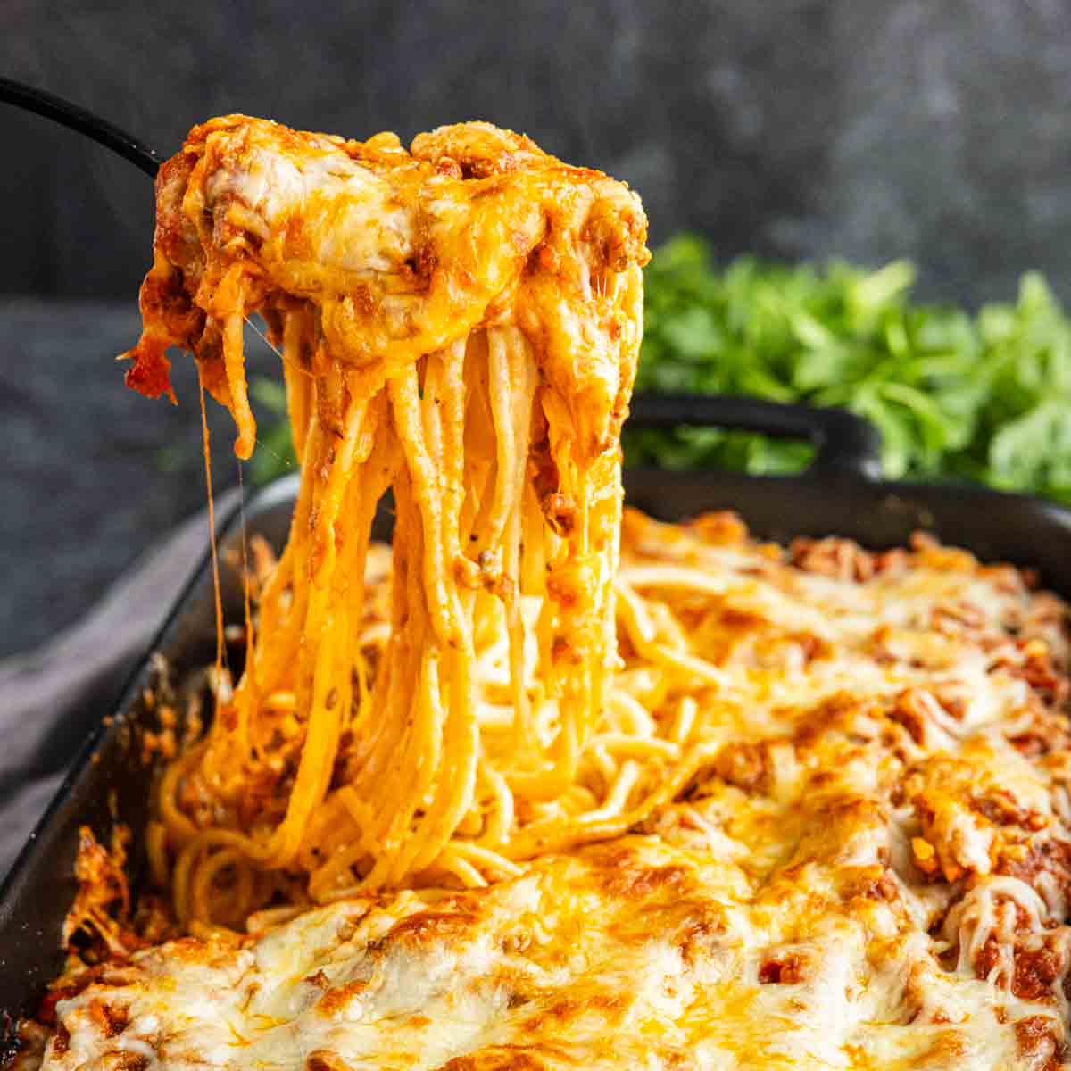 A serving spatula lifts a portion of baked spaghetti topped with melted cheese from a casserole dish, with fresh herbs in the background.