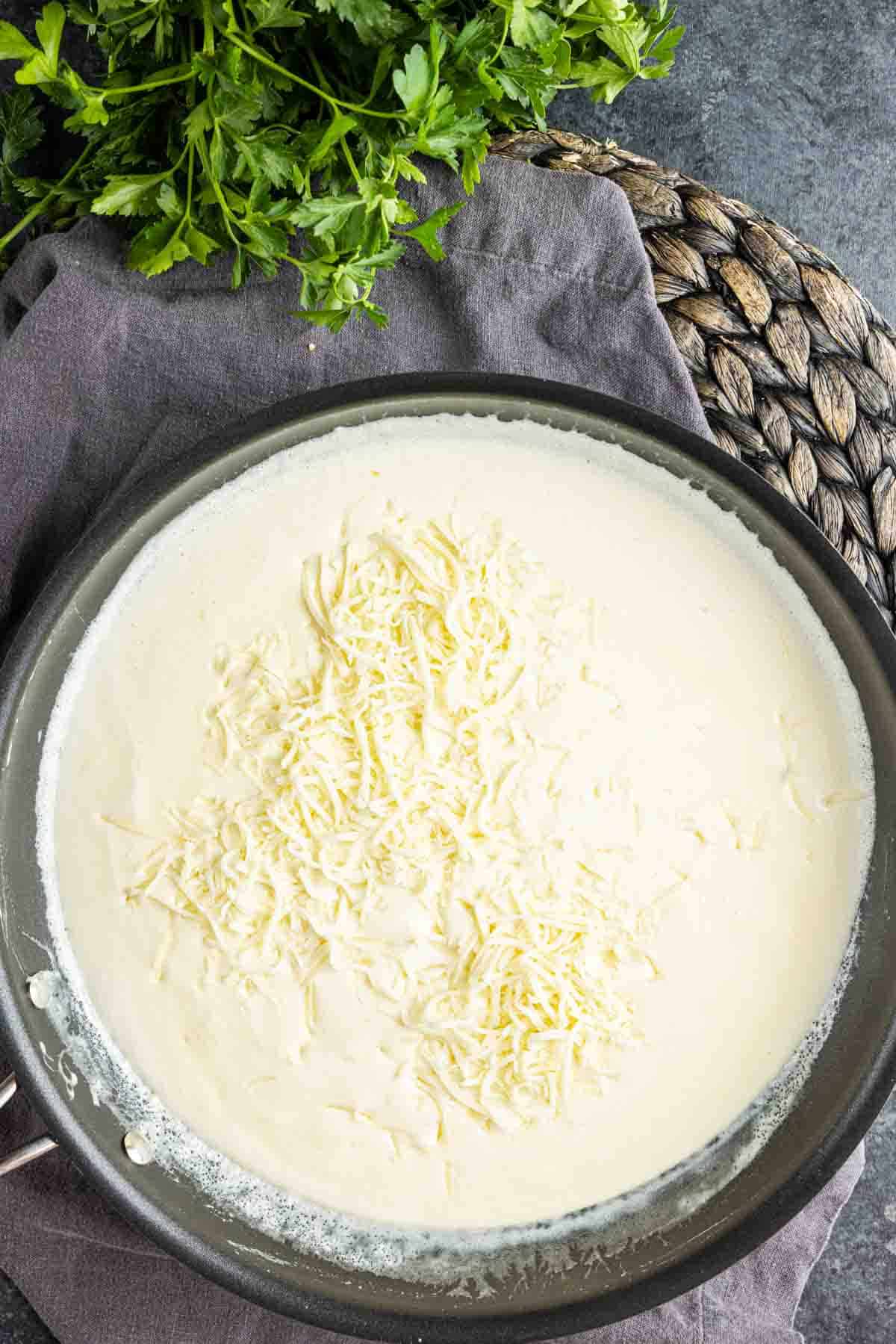 A pan filled with creamy white sauce topped with shredded cheese, placed on a gray cloth next to a bunch of fresh parsley.