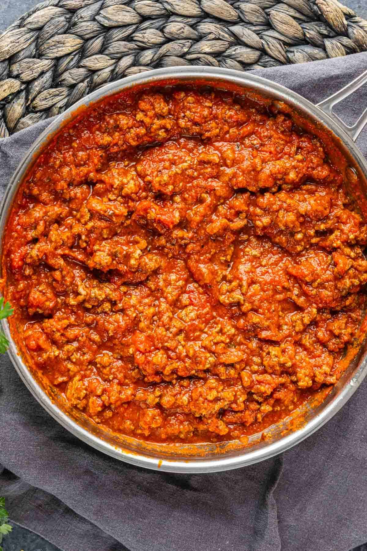 A stainless steel pan filled with cooked ground meat in tomato sauce, resting on a gray cloth with a woven placemat in the background.
