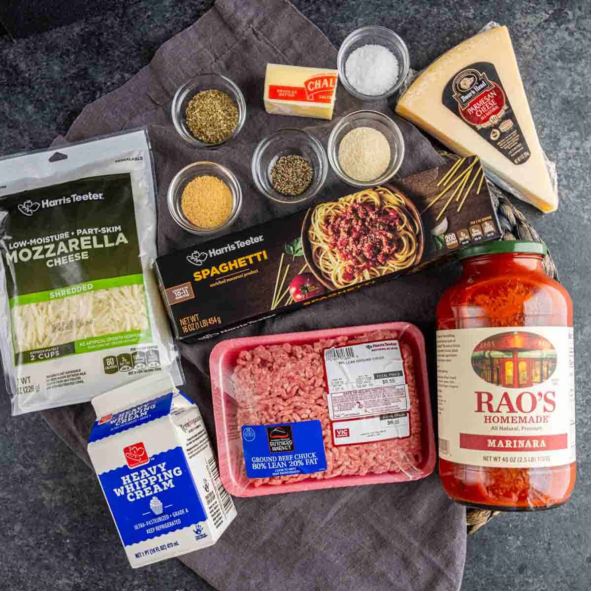 Ingredients for a pasta dish including mozzarella cheese, ground beef, heavy cream, dry spaghetti, marinara sauce, spices, parmesan, and a wedge of Pecorino Romano on a dark countertop.