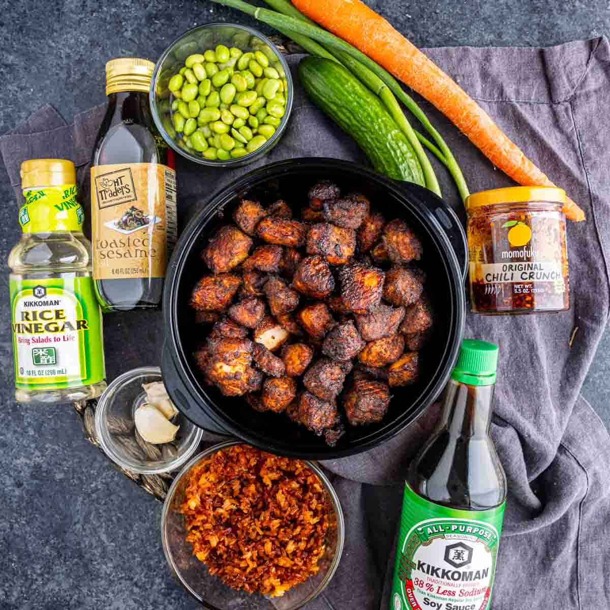 A bowl of cooked, salmon bites surrounded by rice vinegar, sesame oil, edamame, cucumber, carrot, chili crisp dressing, soy sauce, garlic, and crispy rice on a dark surface—perfect for fans of the Salmon Crispy Rice Bowl.