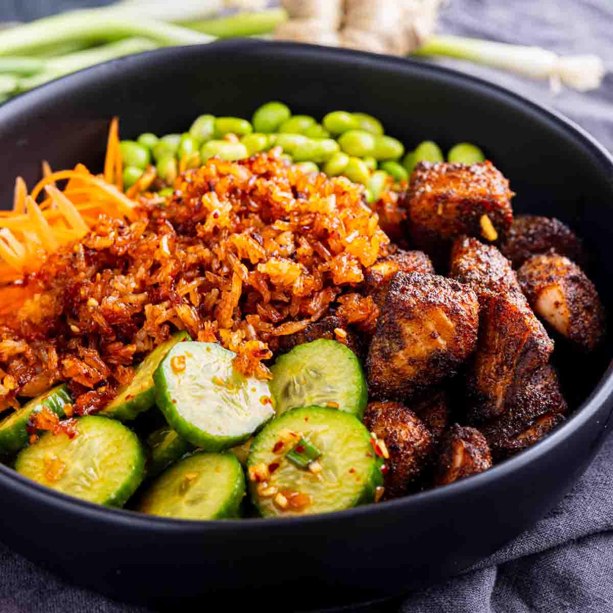 A bowl with sliced cucumbers, shredded carrots, edamame, crispy rice, seasoned salmon bites, and a drizzle of chili crisp dressing.