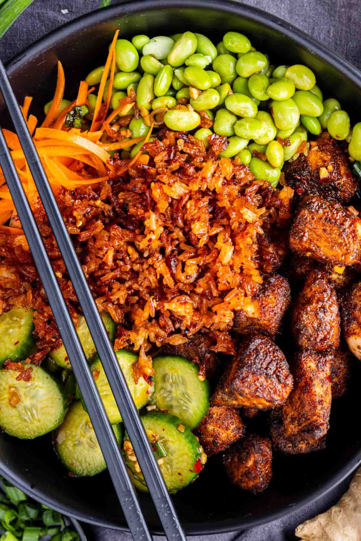 A black bowl with sliced cucumbers, shredded carrots, edamame, seasoned tofu cubes, crispy fried onions, and black chopsticks.